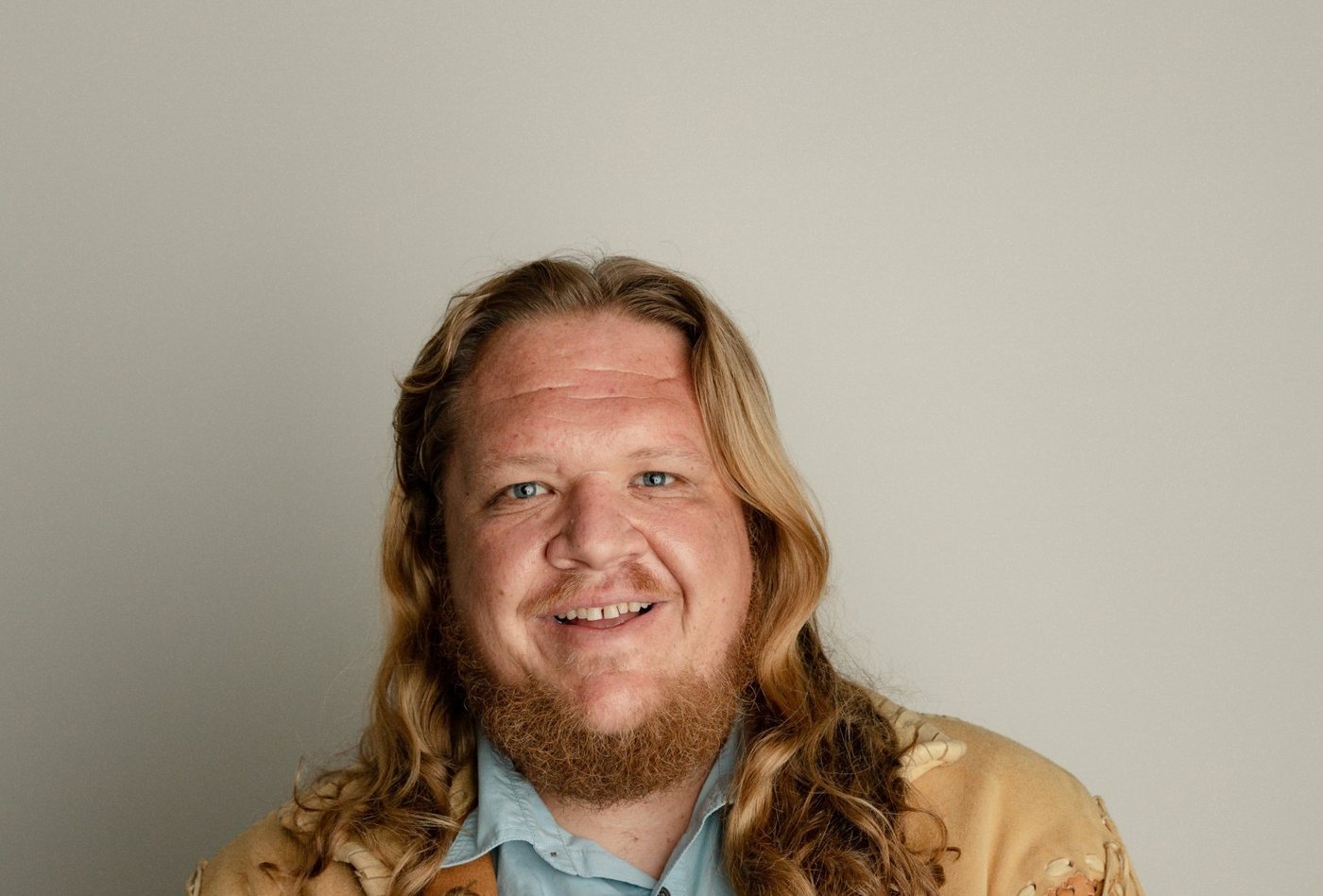Troy, smiling, with long wavy hair and a beard, wearing a fringed leather jacket over a light blue shirt